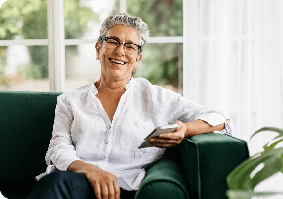 Mulher sorridente de cabelos grisalhos, usando óculos e camisa branca, sentada em um sofá com um celular na mão.